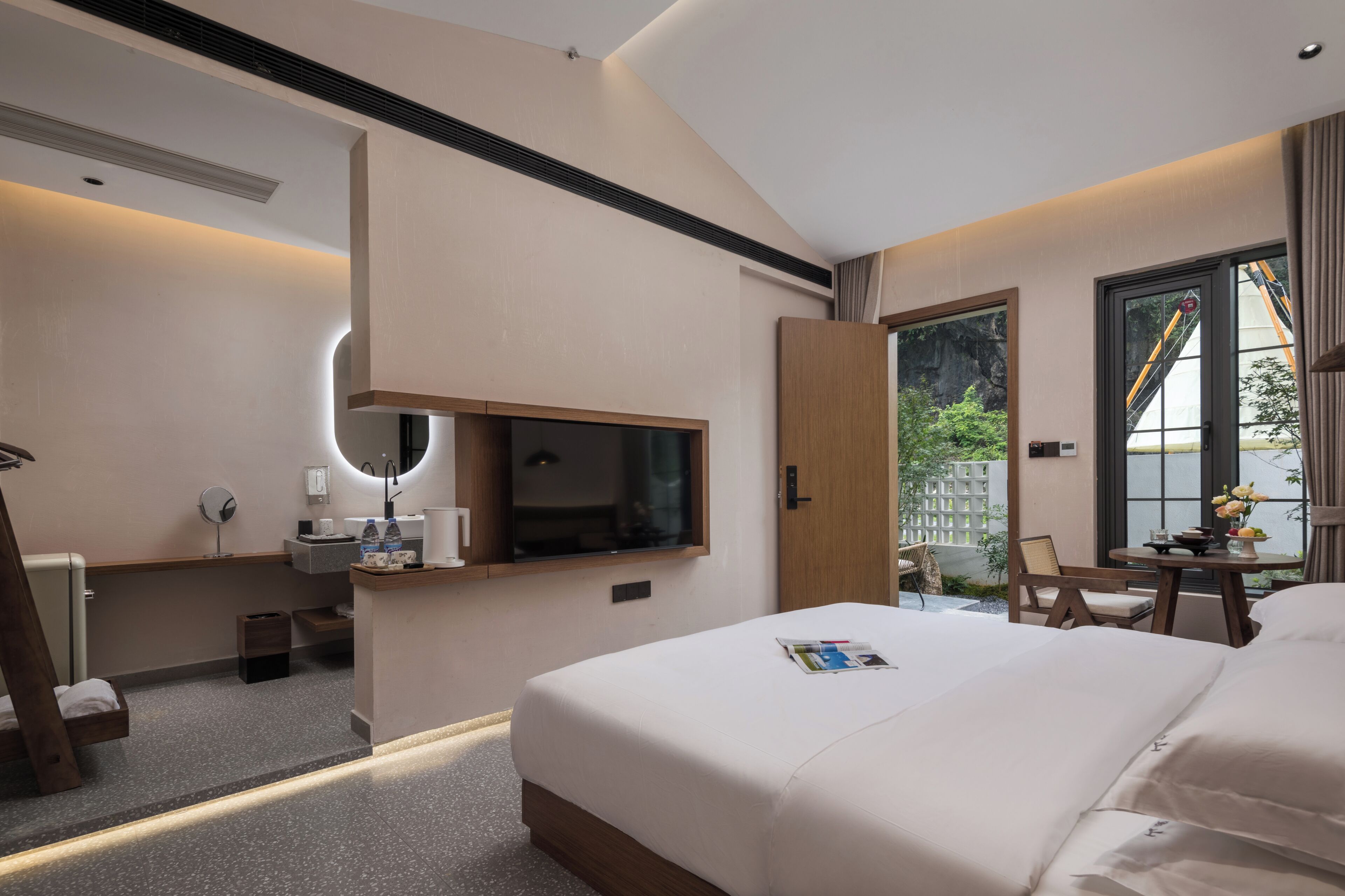 A hotel room with a bed, a TV mounted on the wall, a small desk with a chair, and a window with a view of greenery.