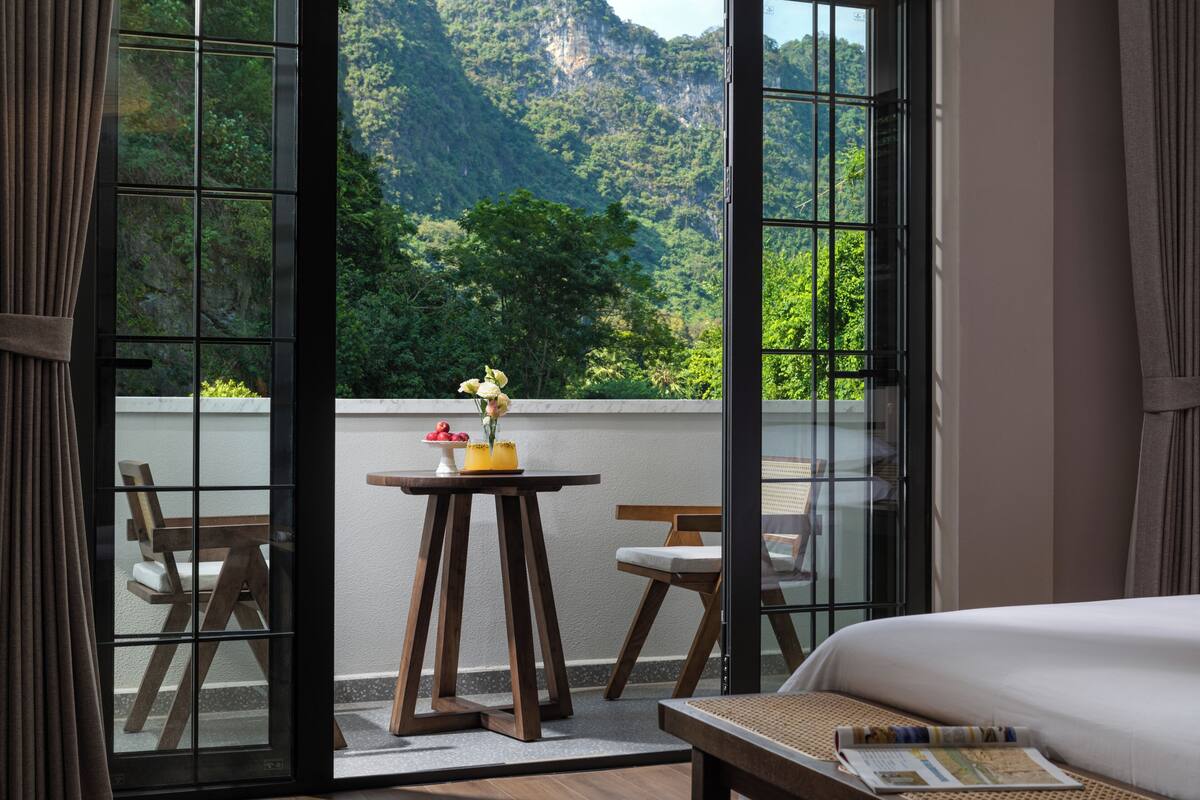 Een hotelkamer met balkon, een bed, een kleintje tafeltje met een vaas en stoelen.