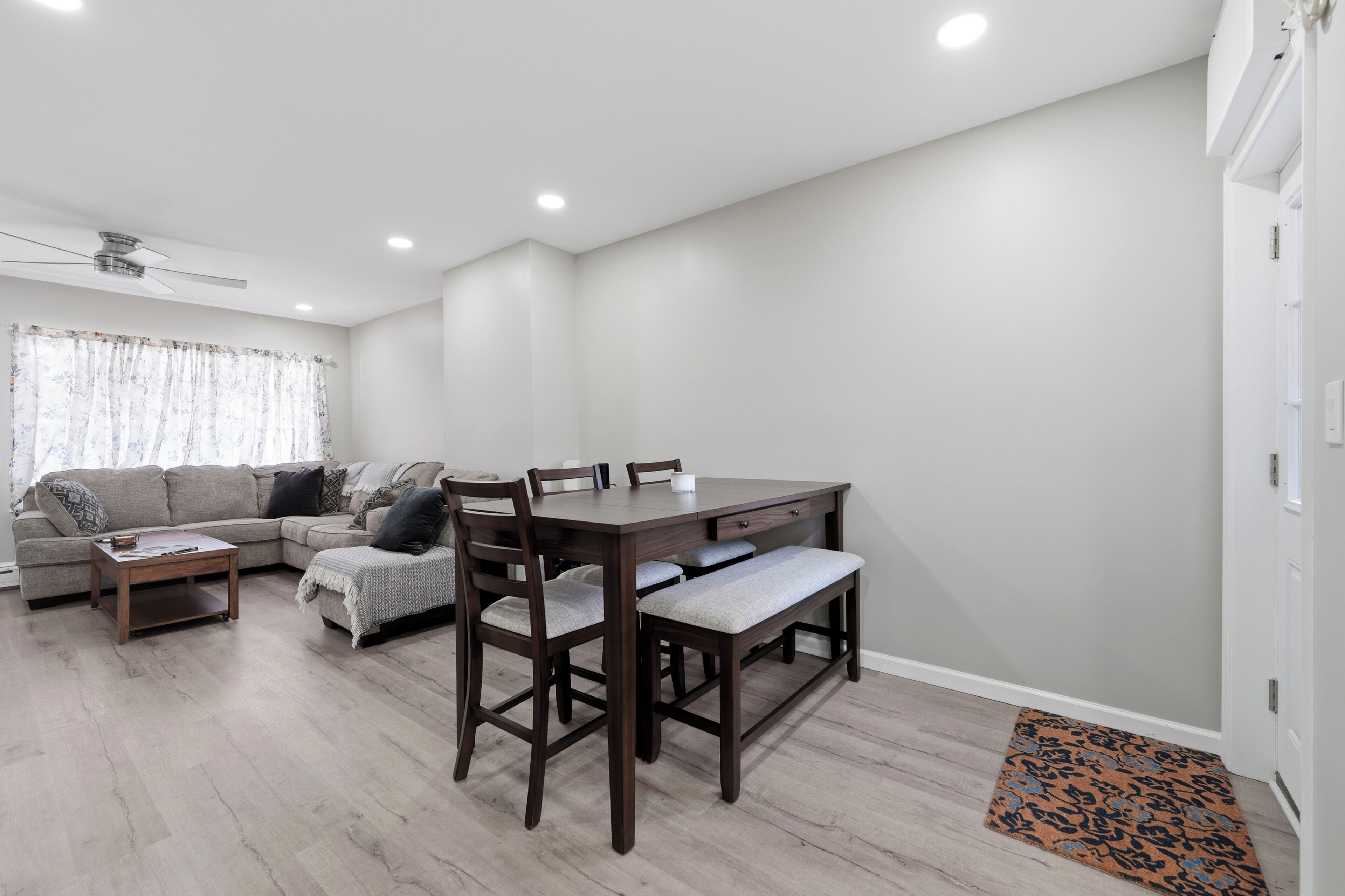 Una sala de estar moderna con un sofá seccional, una mesa de comedor con sillas, un ventilador de techo y una alfombra estampada.