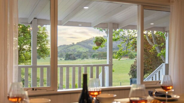 A dining table with wine glasses and a bottle, overlooking a scenic view of a green landscape and hills. - The Meadow Yarra Valley Spa Sauna (Yarra Glen)