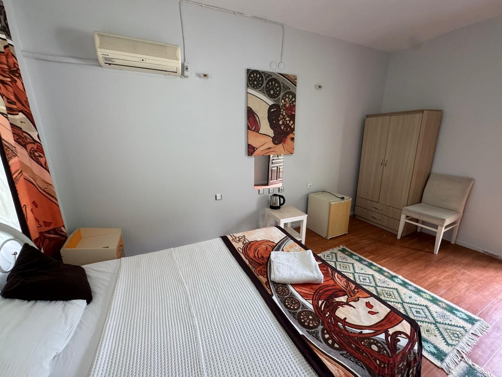A bedroom with a bed, a wardrobe, a chair, a small table, and an air conditioning unit.