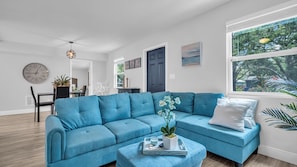 Un salon moderne avec un canapé d’angle bleu, un coin repas agrémenté d’une horloge blanche et une grande fenêtre donnant sur un paysage verdoyant.