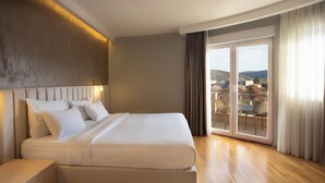 Et moderne hotelværelse med en stor seng, et sengebord, udsigt over byen gennem en skydedør og en balkon med udendørs møbler.