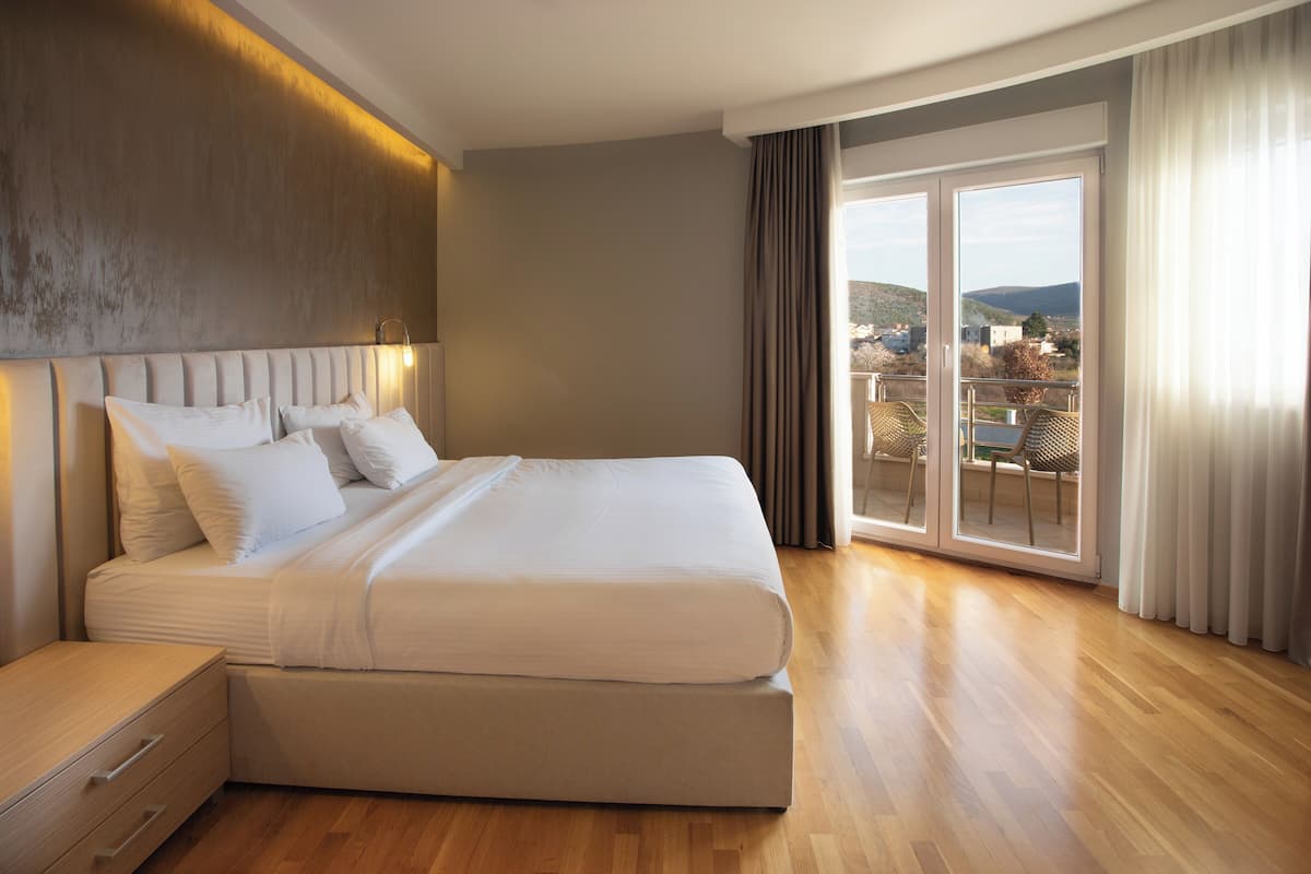 A modern hotel room with a large bed, a bedside table, a view of the city through a sliding glass door, and a balcony with outdoor furniture.