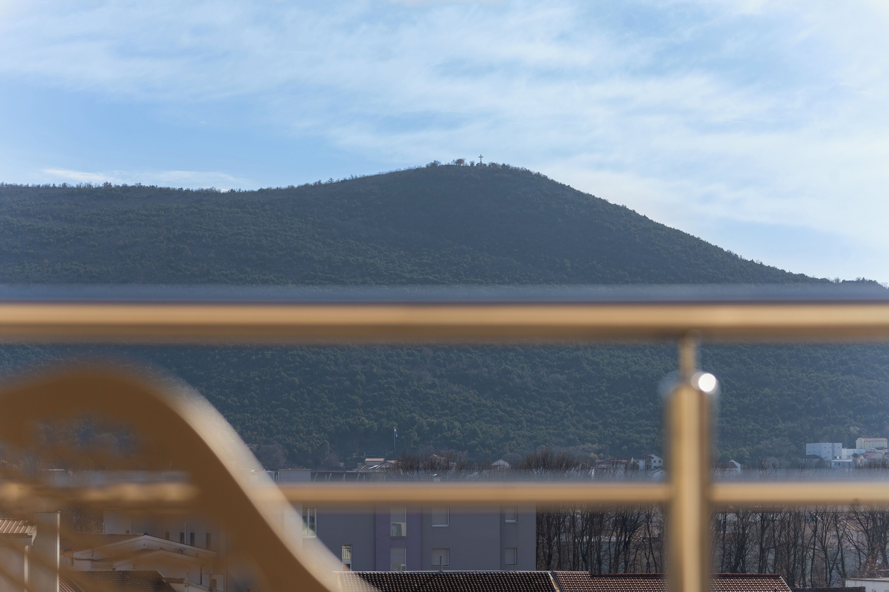 A view of a hill with a cross on top, a clear sky, and buildings in the foreground.