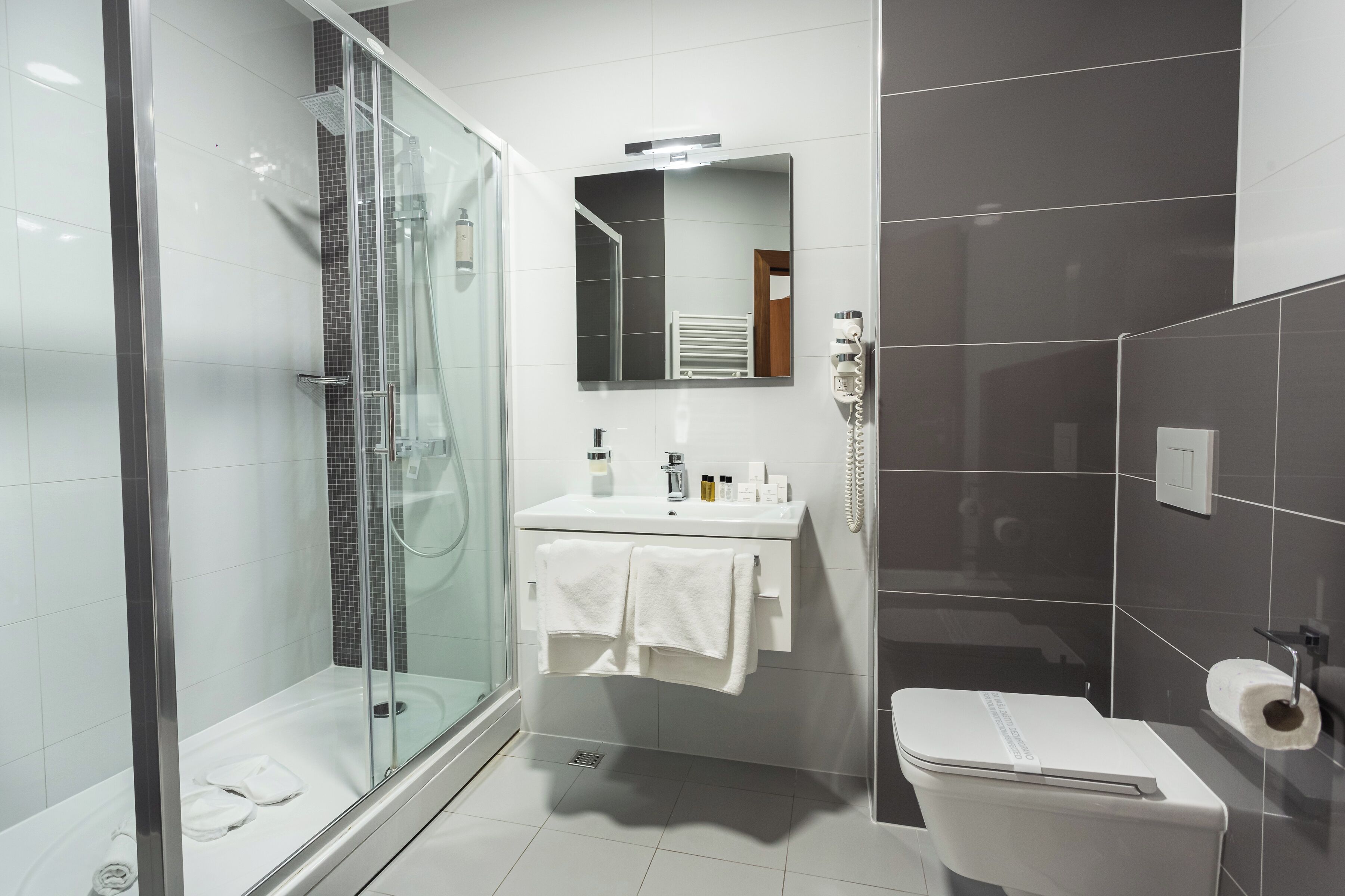 Une salle de bain moderne avec une douche à parois de verre, un lavabo blanc avec un miroir au-dessus et des toilettes dotées d’un porte-serviettes en papier.
