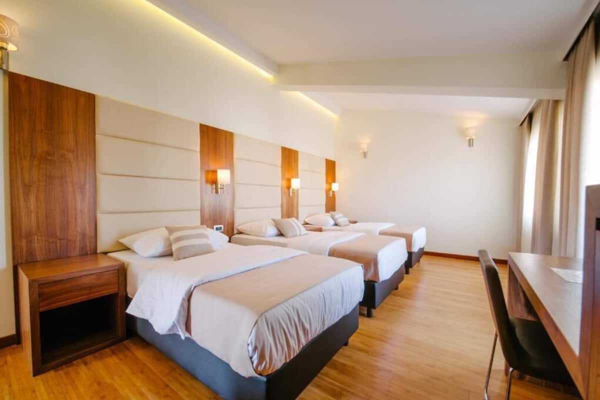A hotel room with three beds, wooden headboards, bedside tables, and a desk with a chair.