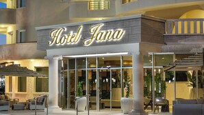 En hotelindgang med et skilt, der siger 'Hotel Jana', udendørs siddepladser med stole og borde, samt en trappe, der fører til indgangen.