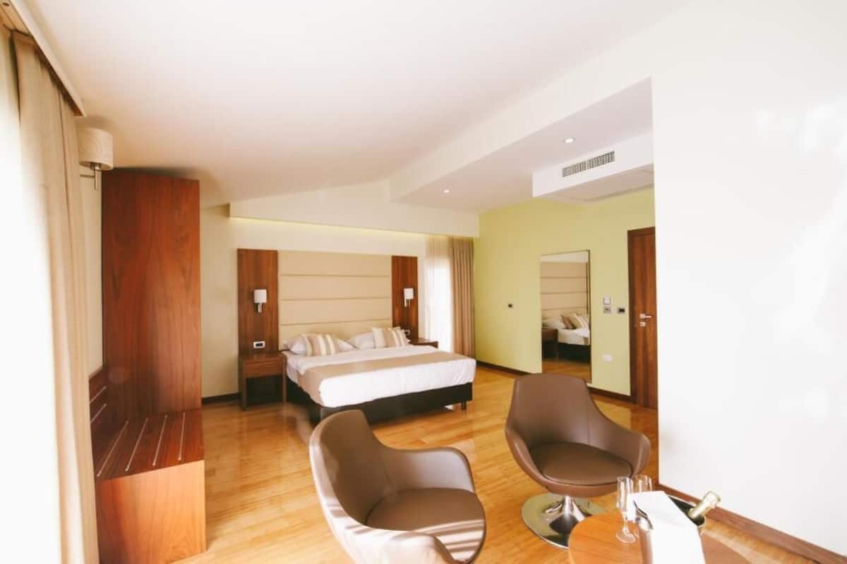 A hotel room with a bed, two armchairs, a small table with a bottle, and a mirror on the wall.