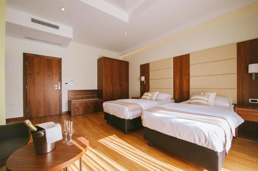 A hotel room with a wooden floor, two beds, a wooden wardrobe, a bedside table, a chair, and a small table with a bottle and glasses.
