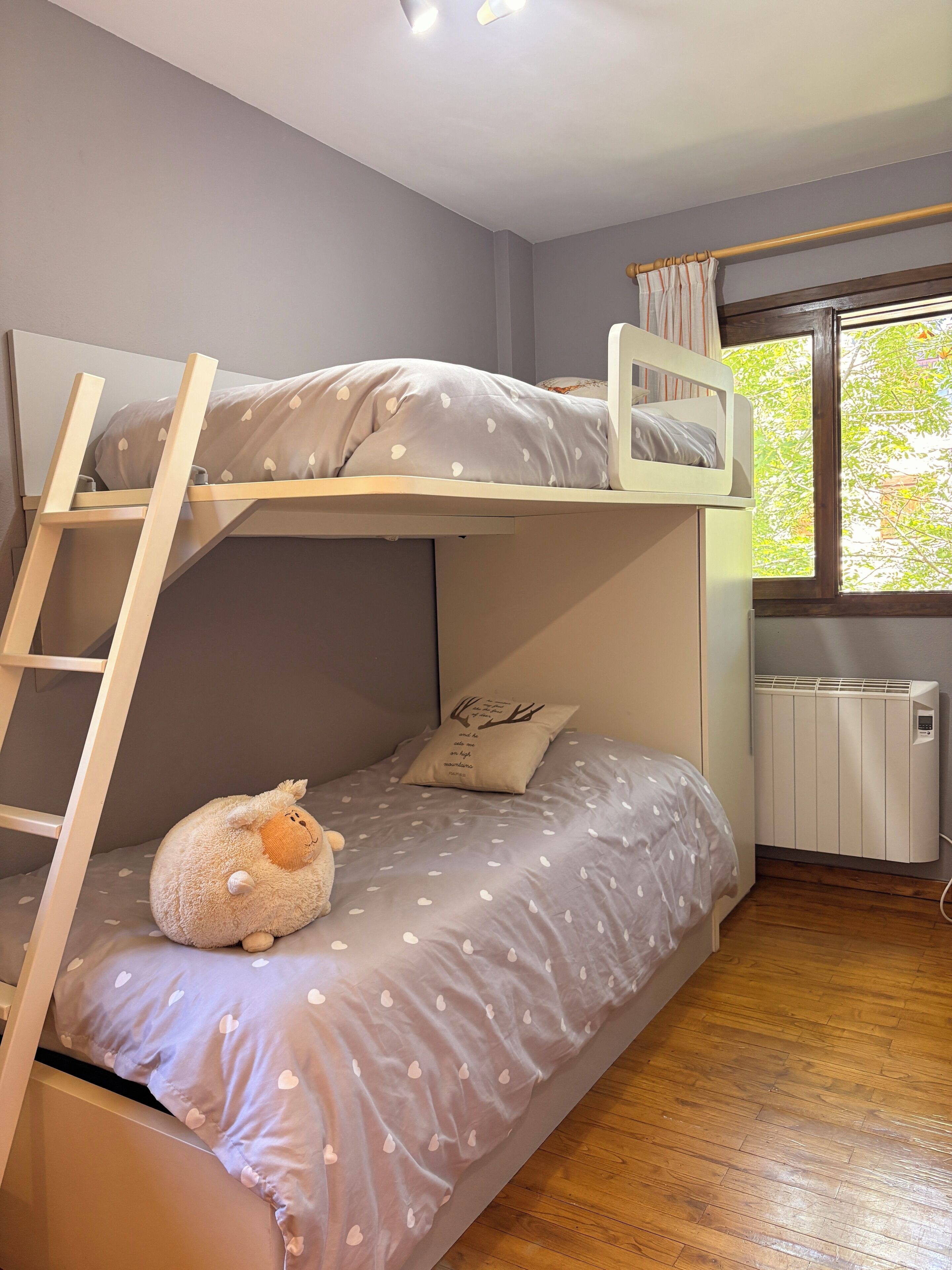 Un lit superposé avec une échelle, un jouet en peluche et une fenêtre donnant sur les arbres.