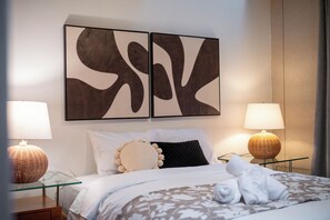 Un lit bien fait, avec une literie blanche, un oreiller noir et un oreiller rond décoratif. Deux tableaux abstraits sont accrochés au mur au-dessus du lit.