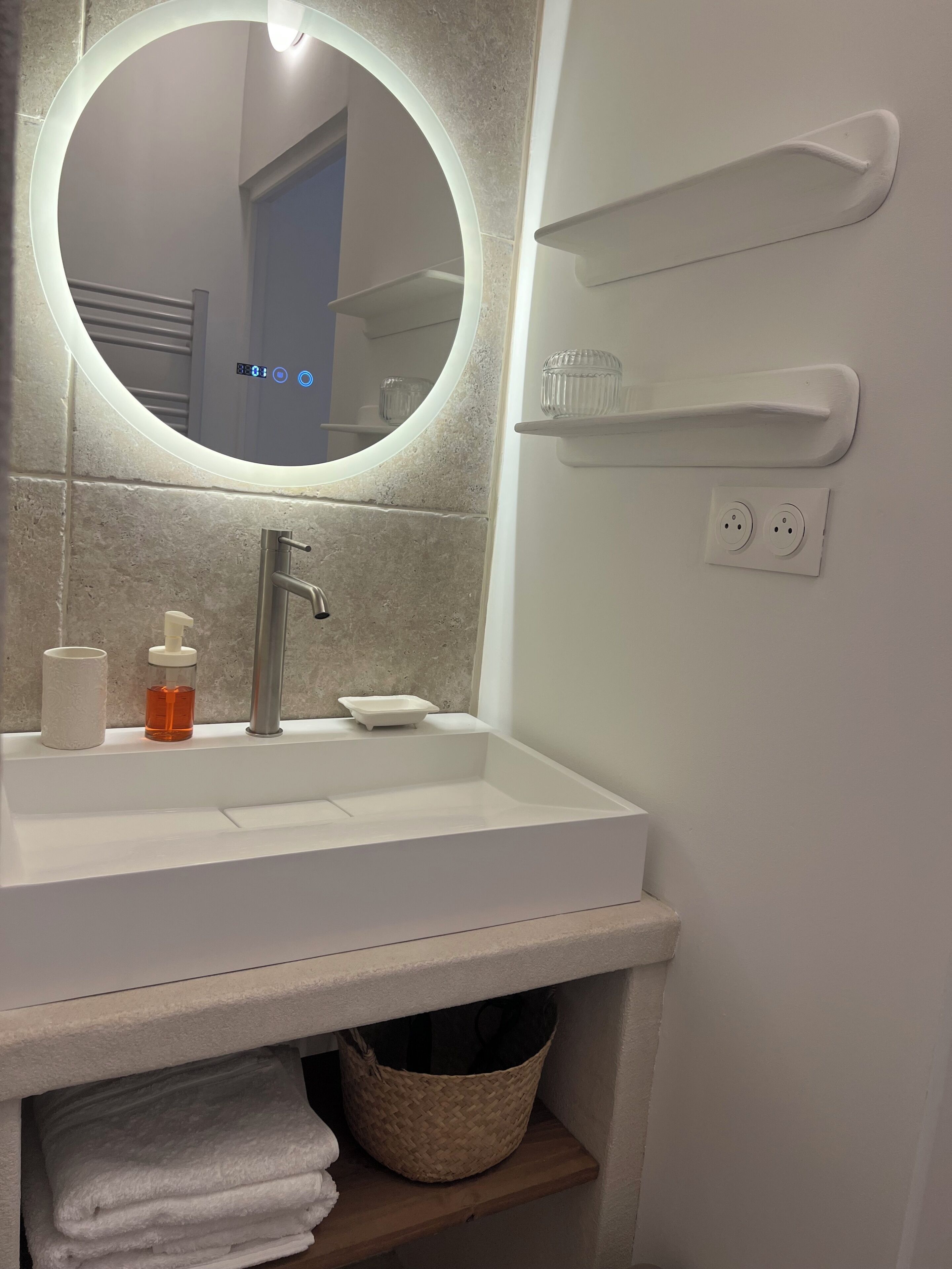 Un baño con un espejo redondo, un lavabo blanco, un dispensador de jabón, un porta cepillos de dientes y un perchero para toallas.