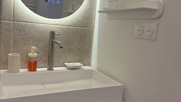 Un baño con un espejo redondo, un lavabo blanco, un dispensador de jabón, un porta cepillos de dientes y un perchero para toallas.