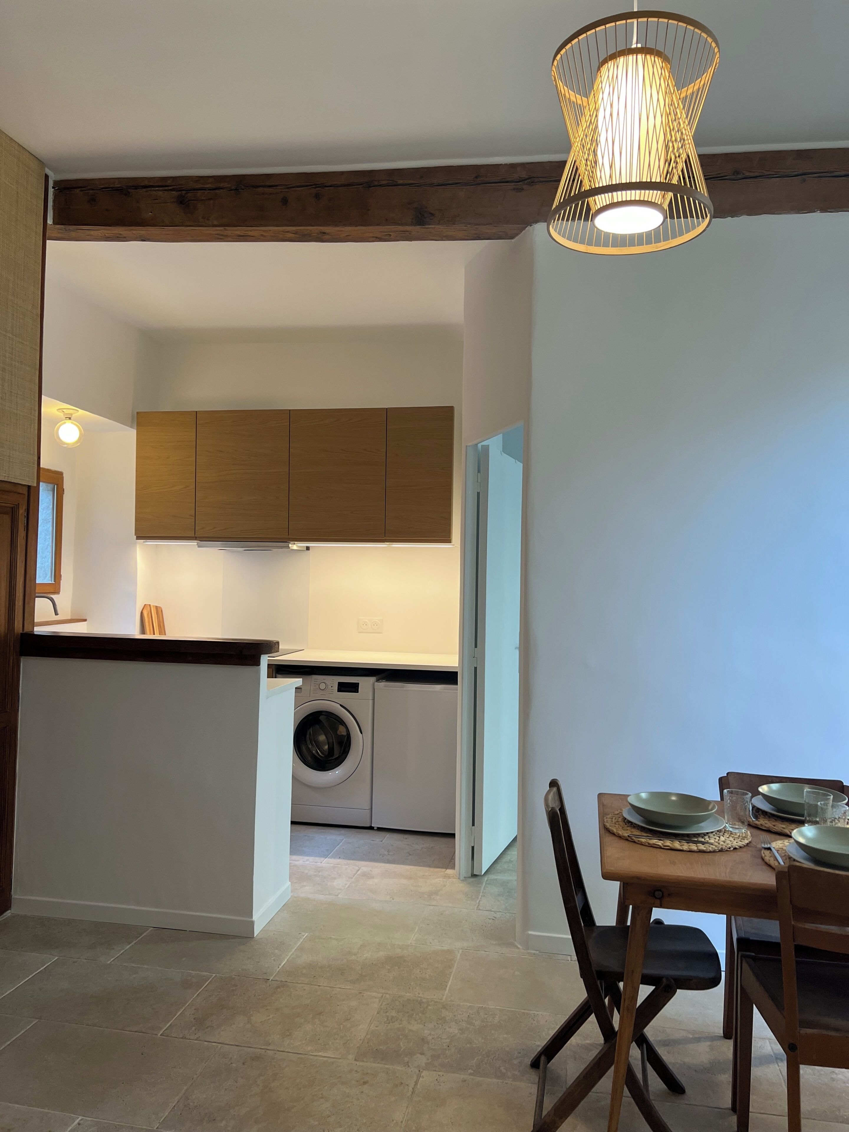 Een kleine keuken met houten kastjes, een wasmachine en een eethoek met een tafel en stoelen.