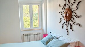 Een slaapkamer met een bed, kussens, een nachtkastje met een lamp, een raam en een decoratie aan de muur.