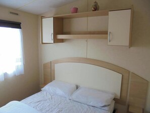 Une chambre à coucher avec un lit, une tête de lit, une fenêtre avec des stores et une étagère sur laquelle se trouvent un bocal et un pot.