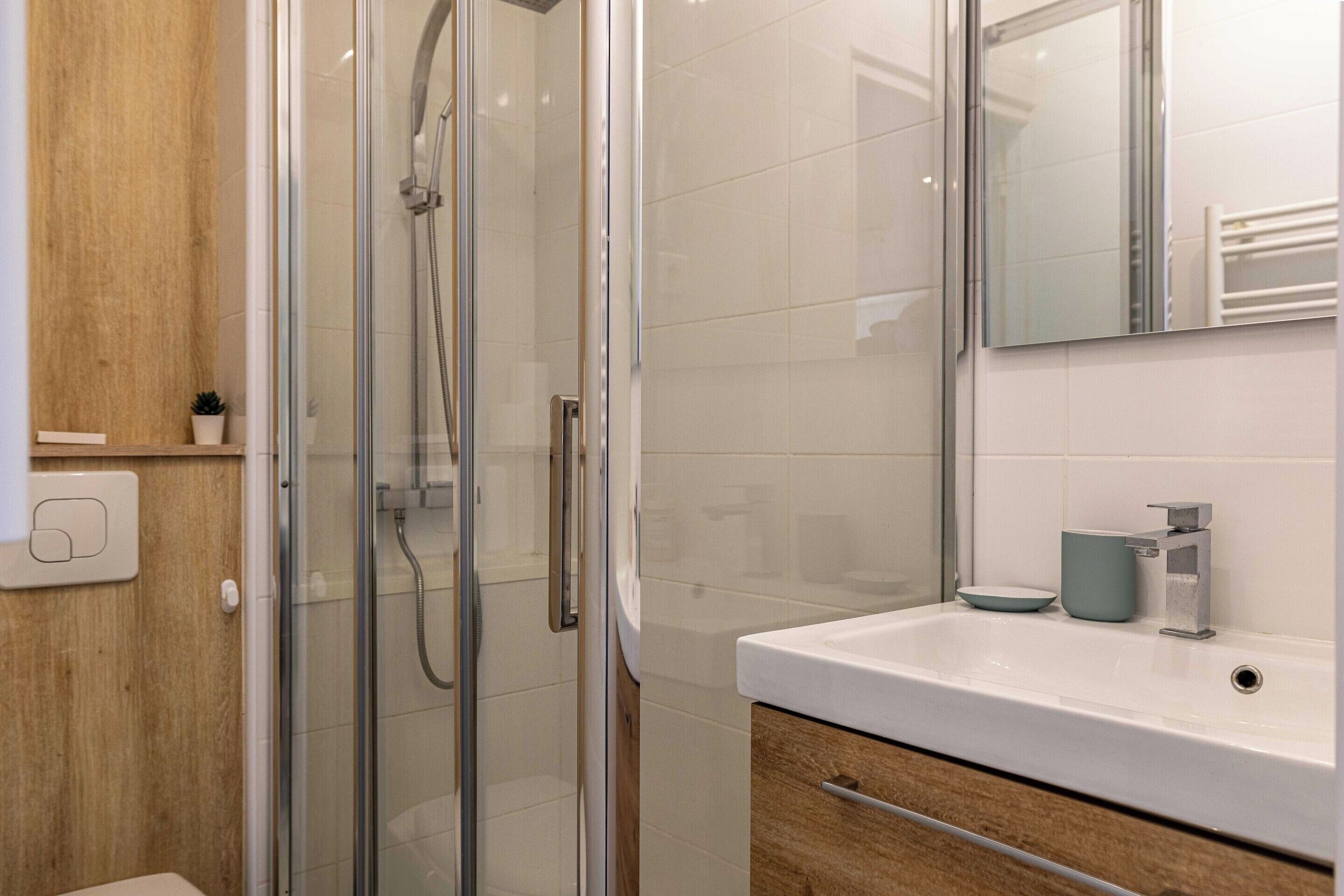 Un baño moderno con cabina de ducha de vidrio, un lavamanos blanco con un jabonera azul y un mueble de tocador de madera.