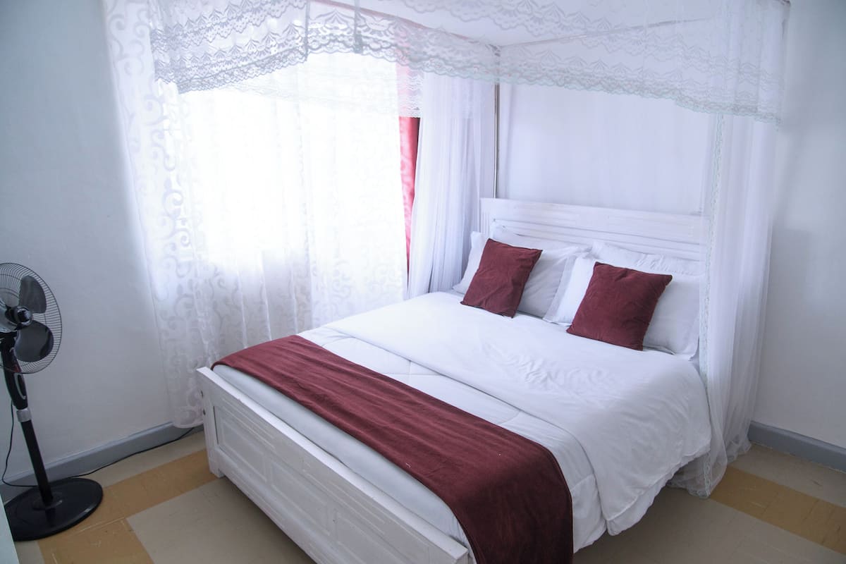 Una cama con ropa de cama blanca y almohadas rojas, un ventilador de pie y cortinas de encaje.