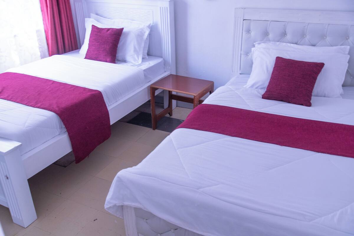 Dos camas individuales con cabeceros blancos, almohadones rojos, una mesita de noche de madera y piso de baldosas.