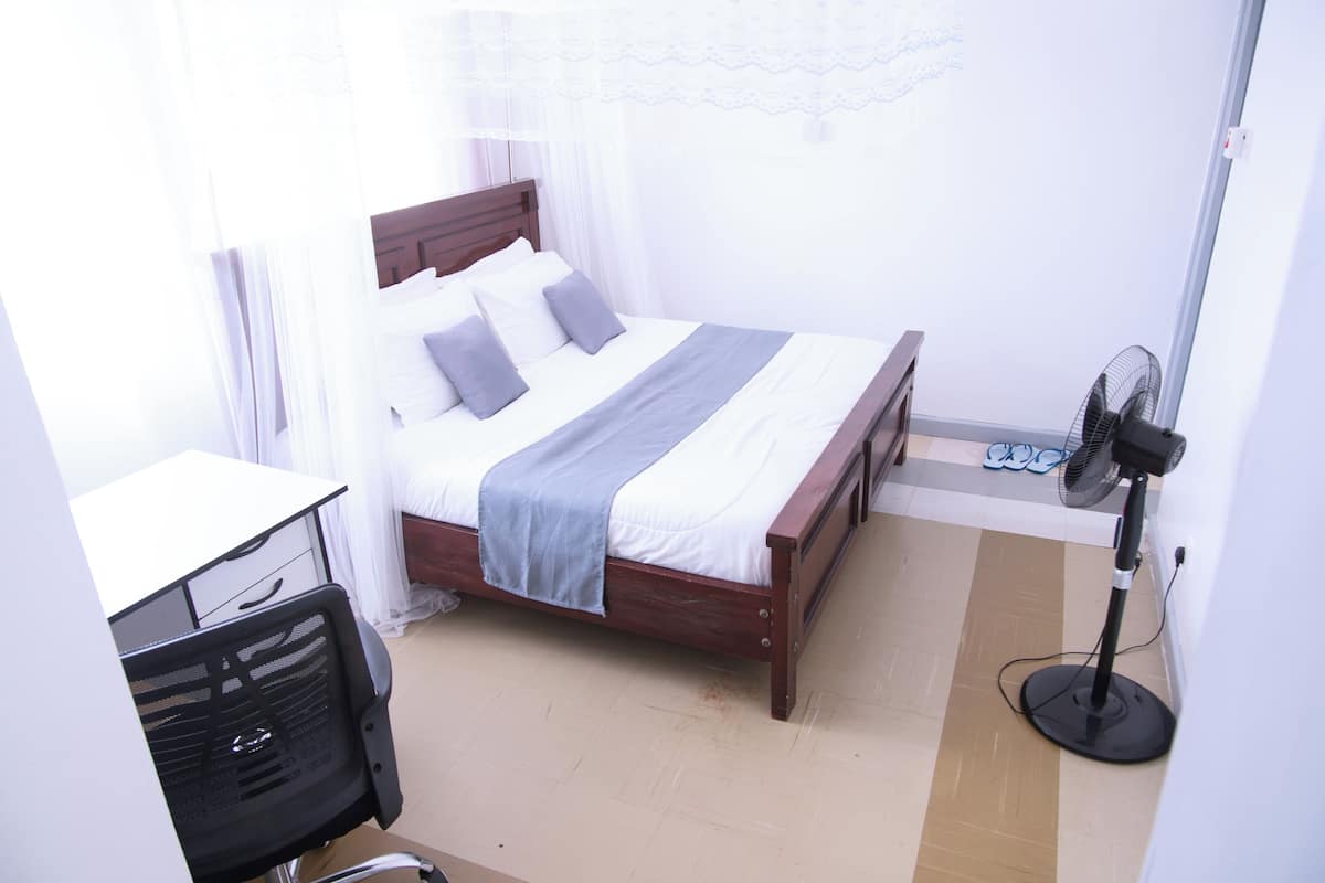 Un dormitorio con cama, escritorio, silla, ventilador y ventana con cortinas.
