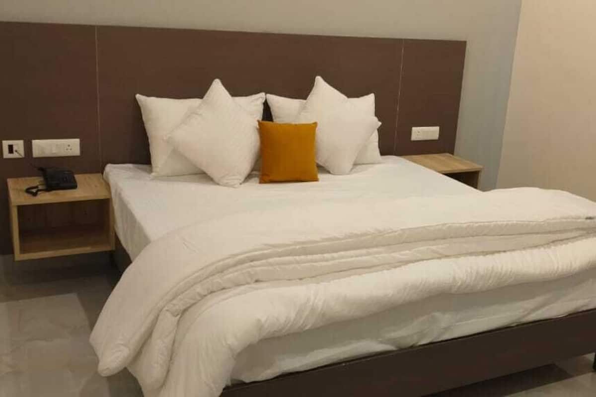 A neatly made bed with white bedding, a brown headboard, and a single orange pillow. There is a wooden nightstand with a phone and a power outlet.
