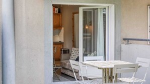 Une petite terrasse avec une table et des chaises, une porte-fenêtre coulissante donnant sur une cuisine et un mur blanc.