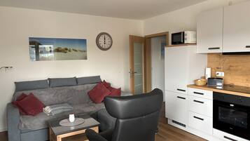 Ein modernes Wohnzimmer mit grauem Sofa, einem schwarzen Sessel, einem kleinen Holztisch, einer Uhr und einer Küchenzeile mit weißen Schränken und einer Mikrowelle.