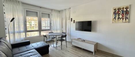 Una sala de estar moderna con un sofá de cuero negro, una mesa de comedor con sillas, un televisor de pantalla plana montado en la pared y una ventana con cortinas traslúcidas.
