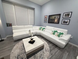 A modern living room with a white sectional sofa, a coffee table, and a large window with blinds. - Modern Unit minutes away from MetLife Stadium (Newark)