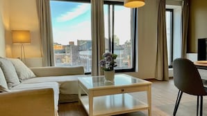 Een moderne woonkamer met een bank, een glazen salontafel, een stoel, een lamp en grote ramen met uitzicht op de stad.