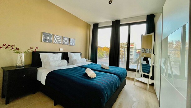 Een hotelkamer met een groot bed, een nachtkastje, een vaas met bloemen en uitzicht op buiten.