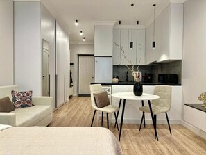 A modern hotel room with a bed, a sofa, a dining table with chairs, a kitchen area, and a hallway leading to another room.