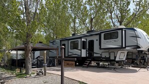 Ein großer Wohnmobil steht auf einem Campingplatz, mit einem überdachten Picknicktisch, einem Grill und einem befestigten Bereich mit Steinen.