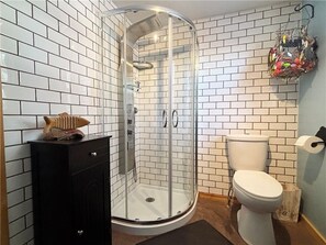 Un baño con paredes de azulejos blancos, ducha con mampara de vidrio, inodoro y un mueble con un objeto decorativo encima.