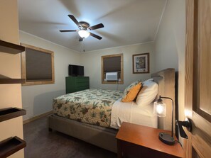 Un dormitorio con cama, ventilador de techo, televisor, ventana con persianas y lámpara de noche.