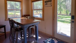 A cabin interior with a wooden dining table, chairs, a 'Welcome to Our Cabin' sign, and large windows offering a view of the forest.