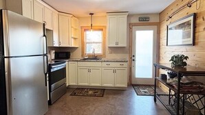 A kitchen with white cabinets, a stainless steel refrigerator, a microwave, and a stove. There is a window with a view of the outdoors, a door with a sign, and a framed picture on the wall.