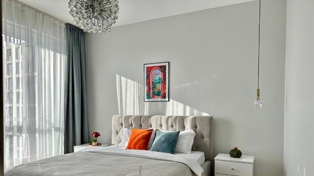 A bedroom with a tufted headboard, a bed with striped bedding, a bedside table, a nightstand, a decorative wall piece, and a hanging light fixture. - BSKYLINE bright 2-rooms apartment at vibrant area (Almaty)