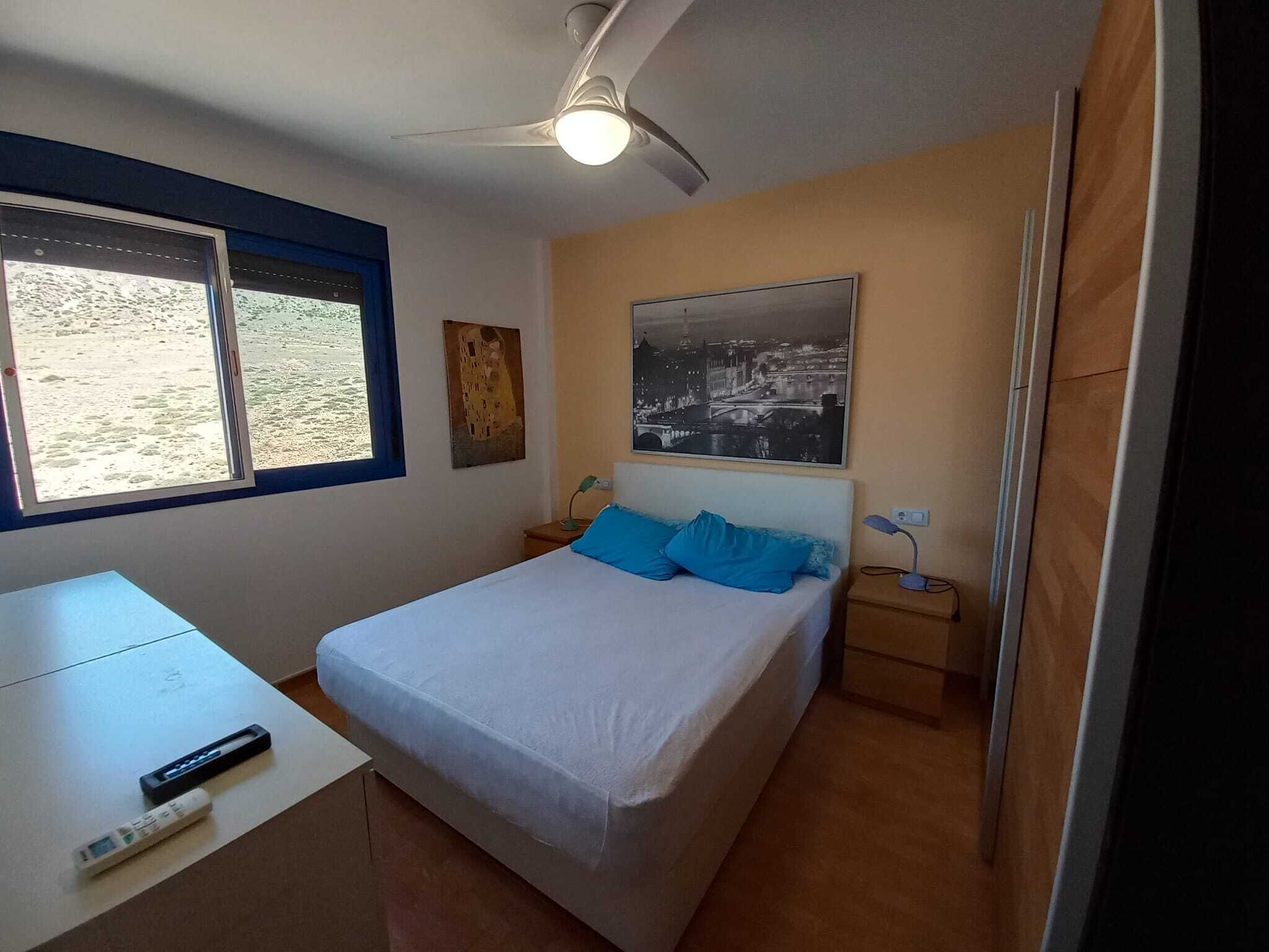 Un dormitorio con cama, escritorio, silla, ventilador de techo, ventana y un cuadro colgado en la pared.