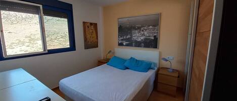Un dormitorio con cama, escritorio, silla, ventilador de techo, ventana y un cuadro colgado en la pared.