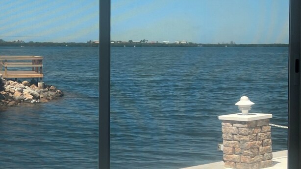 A view through a sliding glass door of a waterfront property with a wooden dock, a stone pillar with a light fixture, and a clear blue sky. - Cozy 2-bedroom home in beautiful Palmetto with spectacular waterfront views. (Palmetto)