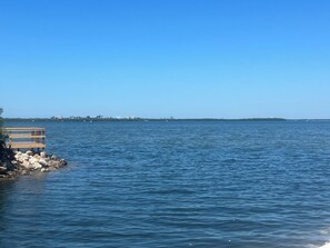 Een houten steiger strekt zich uit over kalm, blauw water, met een heldere lucht en in de verte land met gebouwen.