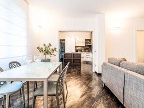 Eine moderne Küche mit weißen Schränken, ein Essbereich mit einem Glastisch und Stühlen sowie ein graues Sofa.