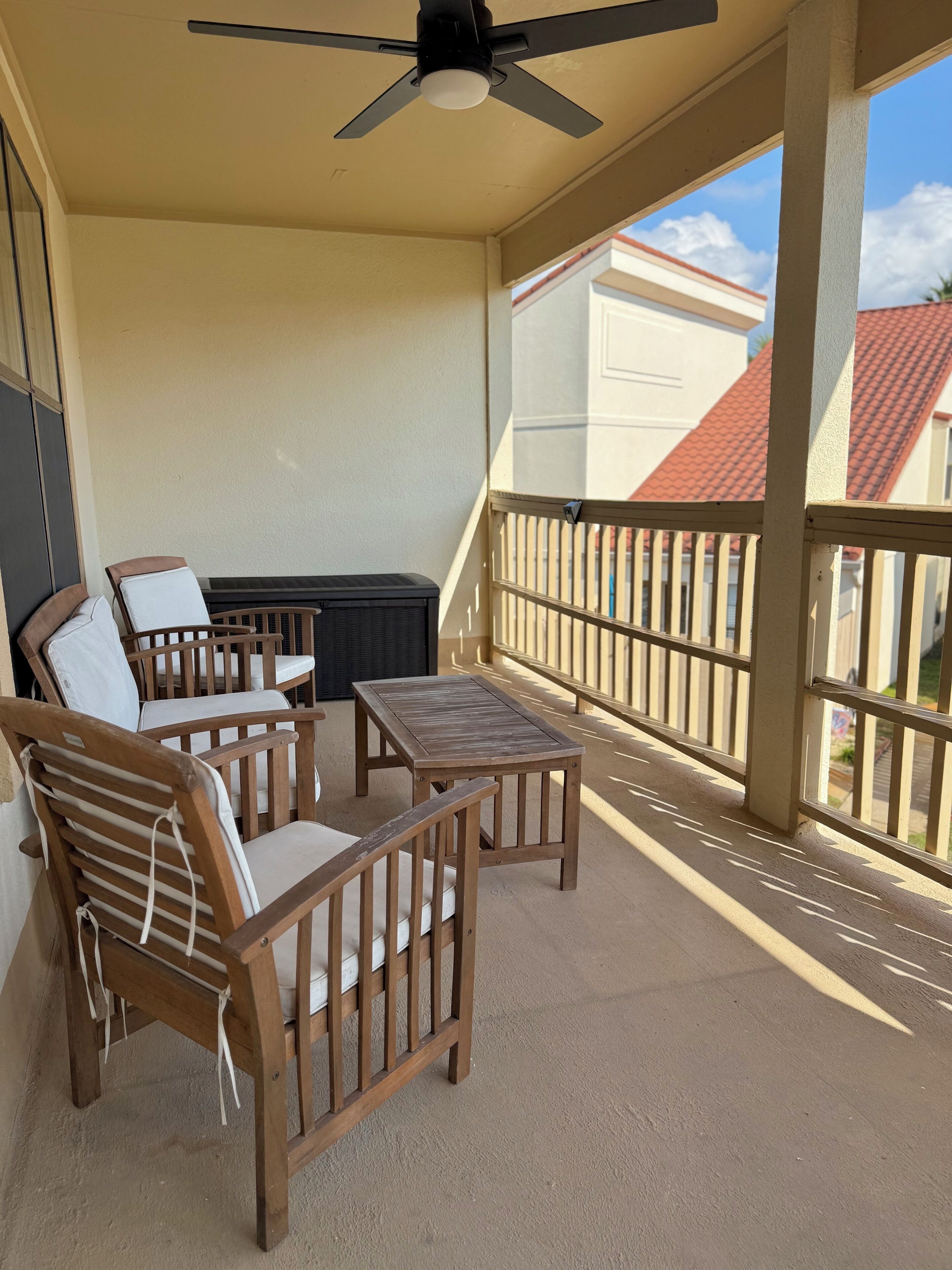 A covered outdoor seating area with a ceiling fan, wooden furniture, and a view of a building with a red roof.