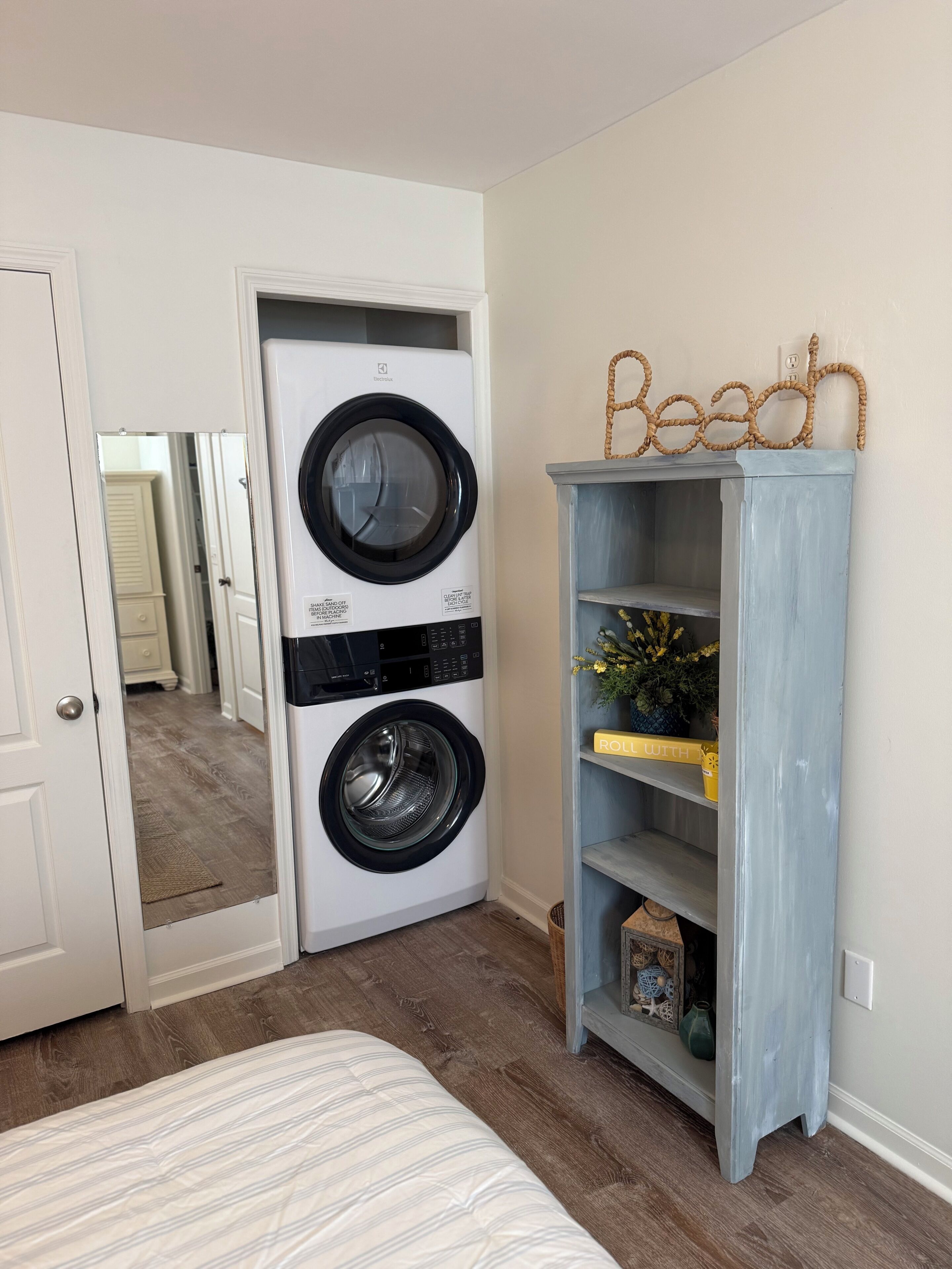 A laundry room with a front-loading washer and dryer, a wooden shelf with decorative items, and a door leading to another room.