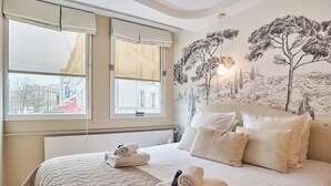 Un dormitorio con una cama grande, dos ventanas con persianas, un radiador y un mural de árboles en la pared.