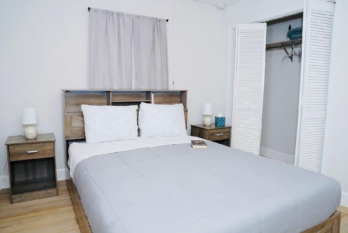 Un dormitorio con cama de madera, ropa de cama blanca, una mesita de noche con lámpara, una mesilla con un libro y un armario con contraventanas blancas.