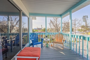 A covered outdoor area with a wooden deck, blue and wooden chairs, a small table with a lantern, and a view of buildings and trees in the background.
