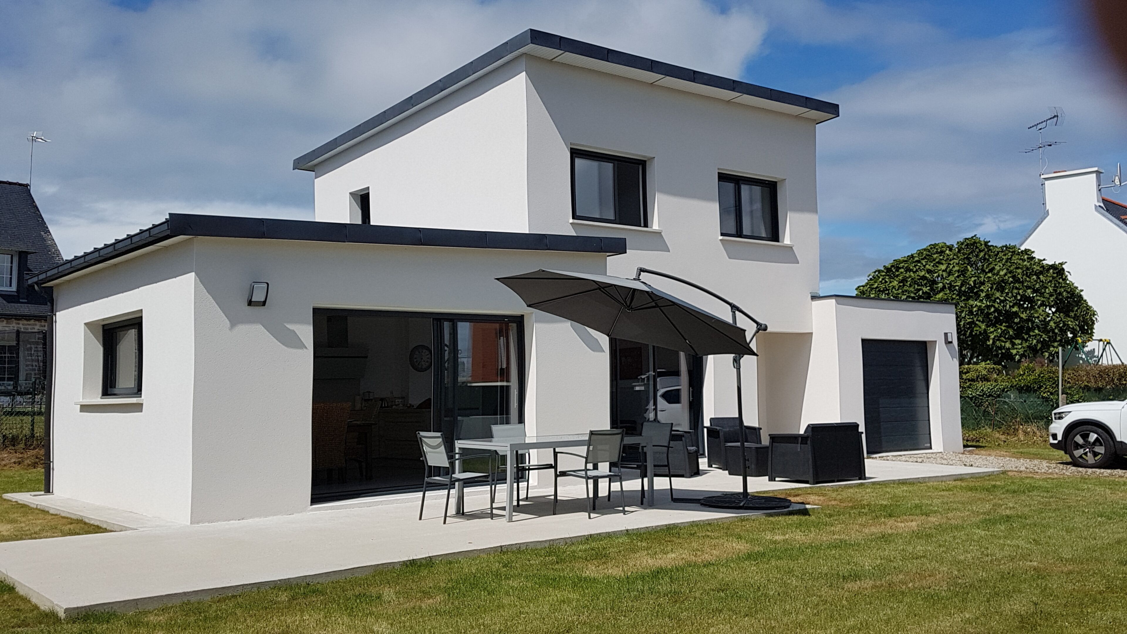 Et moderne hus med to etager, en terrasse, store vinduer og en bil parkeret i nærheden.